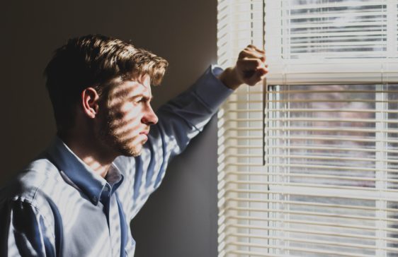 Man looks out window through partially open slats of blinds.